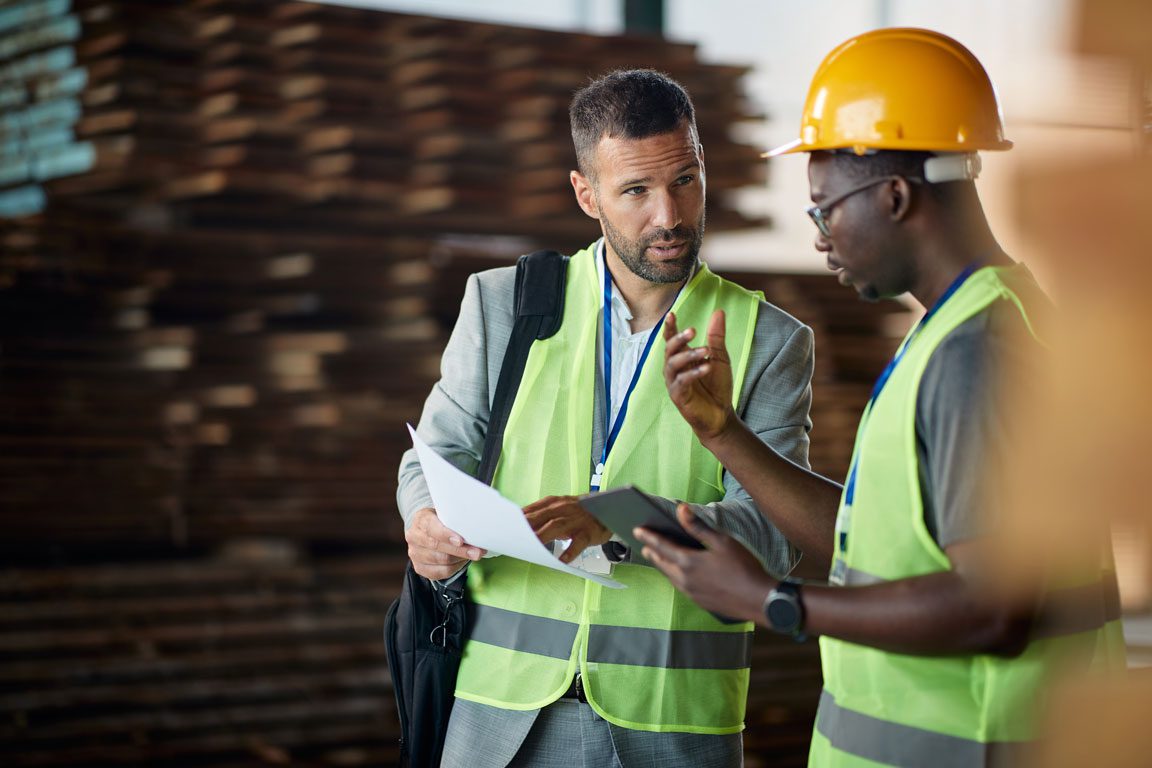 an African American male in a hardhat and a white male with a briefcase both at work in a lumber yard